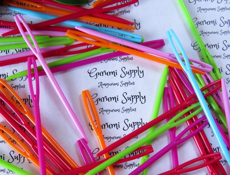 May include: A close-up of a pile of colorful plastic yarn needles. The needles are arranged in a random pattern and are of various lengths. The needles are all the same size and have a small eye at one end. The needles are used for crocheting and knitting.