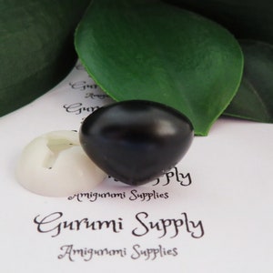 May include: A black plastic safety eye for amigurumi projects. The eye is round and has a glossy finish. The eye is sitting on a white background with the text "Gurumi Supply" printed on it.