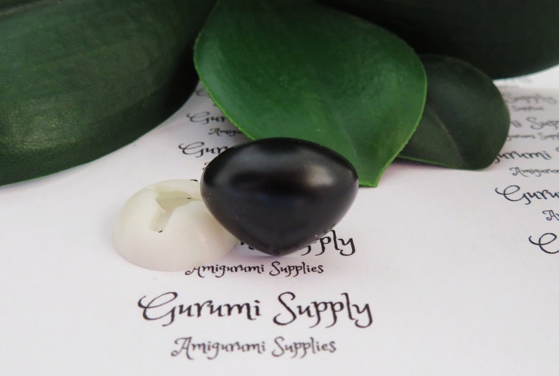 May include: A black plastic safety eye for amigurumi projects. The eye is round and has a glossy finish. The eye is sitting on a white background with the text "Gurumi Supply" printed on it.