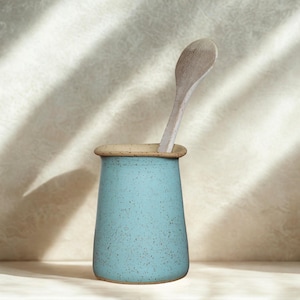 Op de afbeelding: Een turquoise keramische keukengerei houder met een gespikkelde afwerking en een lichtbruine rand, met een houten lepel. De houder is cilindrisch en staat tegen een neutrale achtergrond met schaduwen.