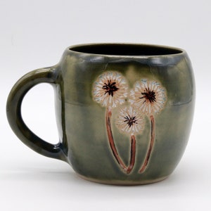 Può includere: Una tazza in ceramica verde con una glassa bianca e tre disegni di dente di leone marroni e bianchi.