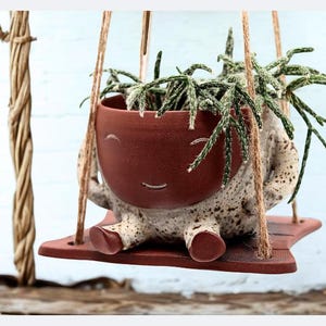 Op de afbeelding: Een grillige keramische plantenpot met een lachend gezicht, hangend aan een houten schommel. De terracotta-kleurige pot bevat een groene vetplant. De pot heeft een gespikkelde, gebroken witte body en is opgehangen met touw.