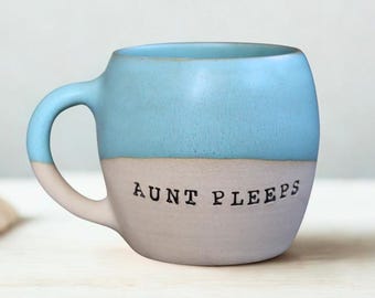 Puede incluir: Una taza de cerámica redondeada con un diseño de dos tonos, con una sección superior azul claro y una sección inferior gris mate. El texto AUNT PEEPS está impreso en negro, en mayúsculas y con una fuente estilo máquina de escribir.