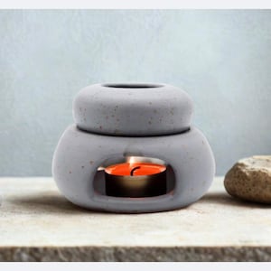 Könnte beinhalten: Ein hellgrauer, gesprenkelter Beton-Ölbrenner mit abgerundetem, zweiteiligem Design und einer zentralen Öffnung, in der eine kleine, runde Teelichtkerze mit orangefarbener Flamme brennt, platziert zwischen zwei dunkelgrauen und einem beigen Stein.