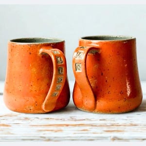 May include: Two orange ceramic mugs with textured surfaces and curved handles. The handles have a decorative design. The mugs are set on a white wooden surface, with a neutral background.