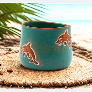 Puede incluir: Una taza de cerámica turquesa con dos ilustraciones de delfines en marrón y blanco. La taza está colocada sobre una estera tejida, con una playa y palmeras al fondo. La taza tiene una forma irregular.