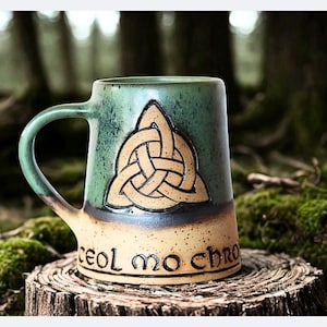 May include: A ceramic mug with a Celtic knot design. The mug has a green top, a tan bottom, and a brown handle. The words "ceol mo chroí" are written on the bottom. The mug is sitting on a tree stump.