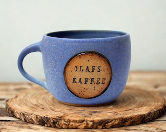 May include: A blue ceramic mug with a speckled finish and a single handle. The front features a circular, tan-colored inset with the text OLAFS KAFFEE stamped in black, uppercase, sans-serif letters across two lines.