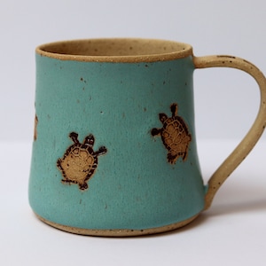 Könnte beinhalten: Eine türkisfarbene Keramiktasse mit braunem Henkel und einem Design aus drei braunen Schildkröten auf der Vorderseite.