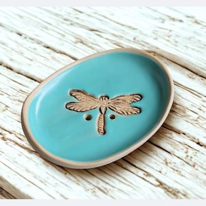 Peut inclure: Un porte-savon en céramique turquoise avec un motif de libellule. Le plat de forme ovale présente une libellule en relief au centre et un bord marron clair. Le plat est posé sur une surface en bois clair.