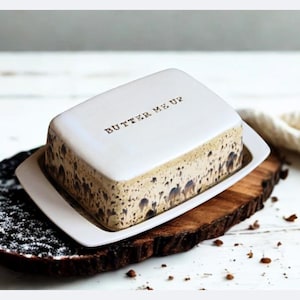 Puede incluir: Una mantequera rectangular de cerámica blanca con el texto "BUTTER ME UP" se encuentra sobre un plato rectangular blanco. La mantequera tiene un diseño moteado marrón y beige. El plato descansa sobre una tabla de madera y un plato decorativo negro.
