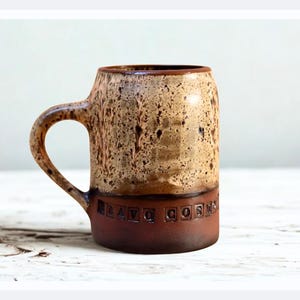 Pode incluir: Uma caneca de cerâmica artesanal com um esmalte salpicado bege e castanho. A caneca tem uma base e uma alça castanho-escuras, com as palavras "Bravo Corn" em relevo na base. A caneca tem cerca de 13 cm de altura.