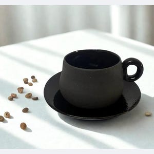Könnte beinhalten: Ein schwarzes Keramik-Kaffeebecher- und Untertassen-Set. Die Tasse hat eine abgerundete Form mit einem kreisförmigen Griff. Die Untertasse ist rund und flach. Kaffeebohnen sind um das Set herum auf einer weißen Oberfläche verstreut.