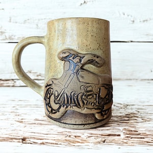 Peut inclure: Une tasse en céramique beige avec un motif de moto. La tasse a une grande anse et une surface texturée. La moto est en relief sur le devant de la tasse, de couleur marron foncé.
