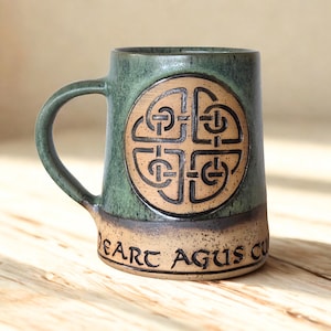 Peut inclure: Une tasse en céramique avec une partie supérieure émaillée verte et une base beige. La tasse présente un motif de nœud celtique circulaire en relief sur le devant et le texte DEART AGUS CU imprimé en noir sur la partie inférieure beige.