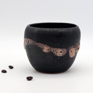 Könnte beinhalten: Eine schwarze Keramiktasse mit gesprenkelter Oberfläche und einem Band aus strukturiertem, hellbraunem Glanz. Drei Kaffeebohnen sind in der Nähe der Tasse verstreut. Die Tasse hat eine abgerundete Form und ein dunkles Inneres.