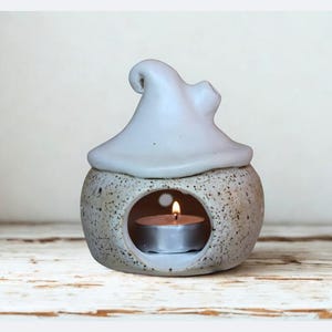 Può includere: Un diffusore di oli essenziali in ceramica con un coperchio a forma di cappello da strega grigio chiaro. La base rotonda e macchiata ha un'apertura circolare con una candela accesa all'interno. Il diffusore è su una superficie di legno chiaro e rustico.