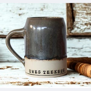 Könnte beinhalten: Ein Bierkrug aus Keramik mit einem grauen oberen Teil und einem beigen unteren Teil. Der Text "UWES TEEKRUG" ist auf dem beigen Teil aufgedruckt. Eine braune Flasche und Holzgegenstände befinden sich im Hintergrund.
