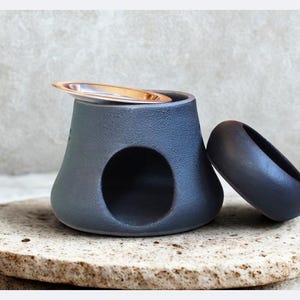 Pode incluir: Um difusor de óleo de cerâmica cinza com uma abertura circular e uma pequena placa de metal na parte superior. Um objeto arredondado cinza escuro repousa ao lado do difusor, tudo sobre uma pedra clara texturizada.