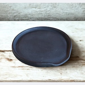 Op de afbeelding: Een donkergrijze keramische lepelhouder met een ovale vorm en een kleine inkeping voor keukengerei. De lepelhouder ligt op een lichtgekleurd houten oppervlak met een grijze achtergrond.