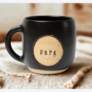 Puede incluir: Taza de cerámica negra con forma redondeada y asa grande. La taza presenta un círculo beige con la palabra "PAPA" impresa en negro. La base de la taza es de color beige claro.