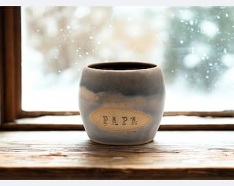 May include: A rounded ceramic mug with a blue and beige glaze, featuring the word PAPA stamped in black capital letters on a lighter, oval-shaped central patch. The mug has a smooth, curved silhouette and a wide opening at the top.