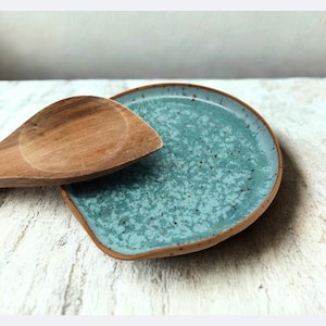 Könnte beinhalten: Ein Holzlöffel liegt auf einer türkisfarbenen Keramik-Löffelablage. Die Löffelablage hat eine gesprenkelte, strukturierte Oberfläche und einen braunen Rand. Der Löffel ist aus hellem Holz.