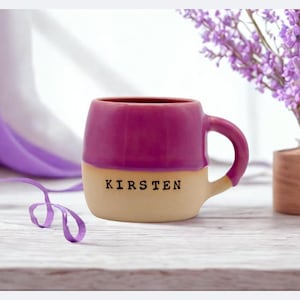 May include: A two-tone ceramic mug with a magenta top and a cream-colored bottom. The name "KIRSTEN" is printed in black letters on the cream section. A small vase with purple flowers sits to the right.