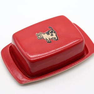 Könnte beinhalten: Rote Keramik-Butterdose mit einem weißen Kuh-Design auf dem Deckel.