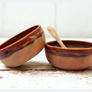 Peut inclure: Deux bols en céramique faits à la main avec un design rustique. Les bols sont bruns avec un bord brun plus foncé et une base brun plus clair. L'un des bols est incliné, et l'autre contient une cuillère en bois.