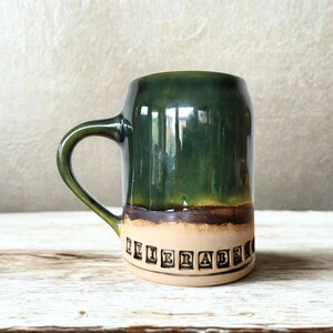 Könnte beinhalten: Handgefertigte Keramiktasse mit dunkelgrüner Glasur im oberen Bereich und beigem Sockel. Die Tasse hat einen gebogenen Henkel und ein braunes Glasurband. Das Wort "FEIERABEND" ist in Schwarz um den Tassenboden gedruckt.