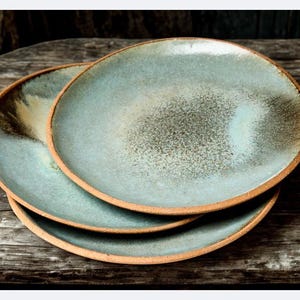 Könnte beinhalten: Drei handgefertigte Keramikplatten, die auf einer Holzoberfläche gestapelt sind. Die Teller haben eine hellblau-grüne Glasur mit gesprenkeltem Aussehen und einen natürlichen braunen Rand. Das rustikale Design verleiht jedem Esszimmer Eleganz.