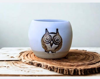May include: A rounded ceramic planter with a smooth white glazed upper section and a natural tan base. The front features a dark brown, etched owl design with large circular eyes, pointed ears, and a small beak, centered on the white surface.