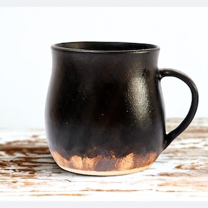 Könnte beinhalten: Dunkelbraune Keramiktasse mit rundem Korpus und Henkel. Die Tasse hat eine strukturierte Oberfläche und einen hellbraunen Streifen am Boden. Die Tasse ist etwa 10 cm hoch, ideal für Kaffee oder Tee.