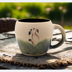 May include: A ceramic mug with a cat silhouette design. The mug has a cream-colored upper half and a green-blue lower half. The interior is black, and the handle is a gradient of green-blue. The mug sits on a wooden table.