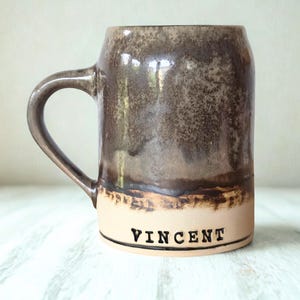 Peut inclure: Une tasse en céramique au design bicolore, avec une glaçure brune tachetée sur la partie supérieure et une base beige clair lisse. Le nom VINCENT est imprimé en lettres capitales noires près du bas de la tasse, dotée d'une anse incurvée.