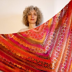 Pode incluir: Um xale triangular de crochê com um gradiente vibrante de cores quentes, incluindo vermelho profundo, laranja brilhante, magenta e amarelo dourado. O tecido exibe padrões rendados complexos, pontos texturizados e uma borla vermelha.