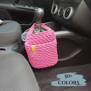 Könnte beinhalten: Eine pinkfarbene gehäkelte Auto-Organizer-Tasche mit kleiner Tasche und Griff, die am Schalthebel eines Autos hängt. Die Tasche hat ein kleines Lederetikett. Das Bild enthält den Text "50+ COLORS".