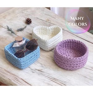 Op de afbeelding: Drie gehaakte manden in verschillende kleuren en vormen. Een lichtblauwe vierkante mand, een witte hartvormige mand en een ronde paarse mand.