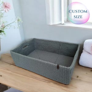 Könnte beinhalten: Ein grauer gehäkelter Aufbewahrungskorb mit einem Ledergriff. Der Korb steht auf einer Holzoberfläche. "CUSTOM SIZE" steht auf einer blauen und violetten Blase im Hintergrund.