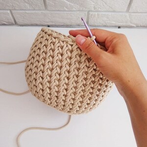 Peut inclure: Gros plan d'un panier en crochet beige en cours de fabrication avec un crochet violet. Le panier est rond et pr&eacute;sente un motif textur&eacute;.