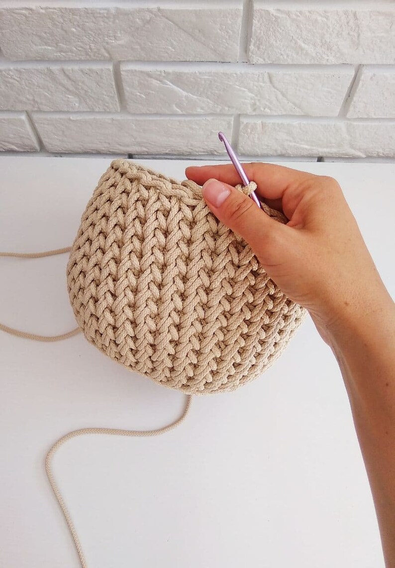 Peut inclure: Gros plan d'un panier en crochet beige en cours de fabrication avec un crochet violet. Le panier est rond et pr&eacute;sente un motif textur&eacute;.