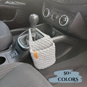 Peut inclure: Un organisateur de voiture en crochet gris avec une poignée, suspendu près du levier de vitesses. L'organisateur a une poche et une petite étiquette en cuir. L'image comprend le texte "50+ COLORS" dans une bannière bleue.