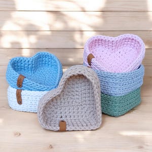 Può includere: Quattro cesti a forma di cuore all'uncinetto in diversi colori: rosa, azzurro, bianco e verde. I cesti sono impilati uno sopra l'altro.