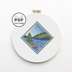 May include: A diamond-shaped cross-stitch pattern featuring a lake scene with a green tree, blue water, and a green hill in the background. There are three birds flying in the sky above the lake.