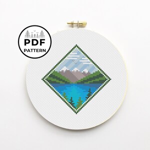 May include: A cross-stitch pattern of a mountain landscape with a lake, trees, and a blue sky. The pattern is in a diamond shape and is framed in a white embroidery hoop.