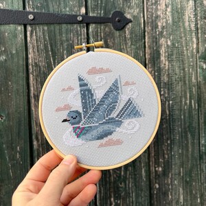 May include: A blue and white cross-stitch embroidery of a stylized bird with pink and white clouds in a wooden embroidery hoop.