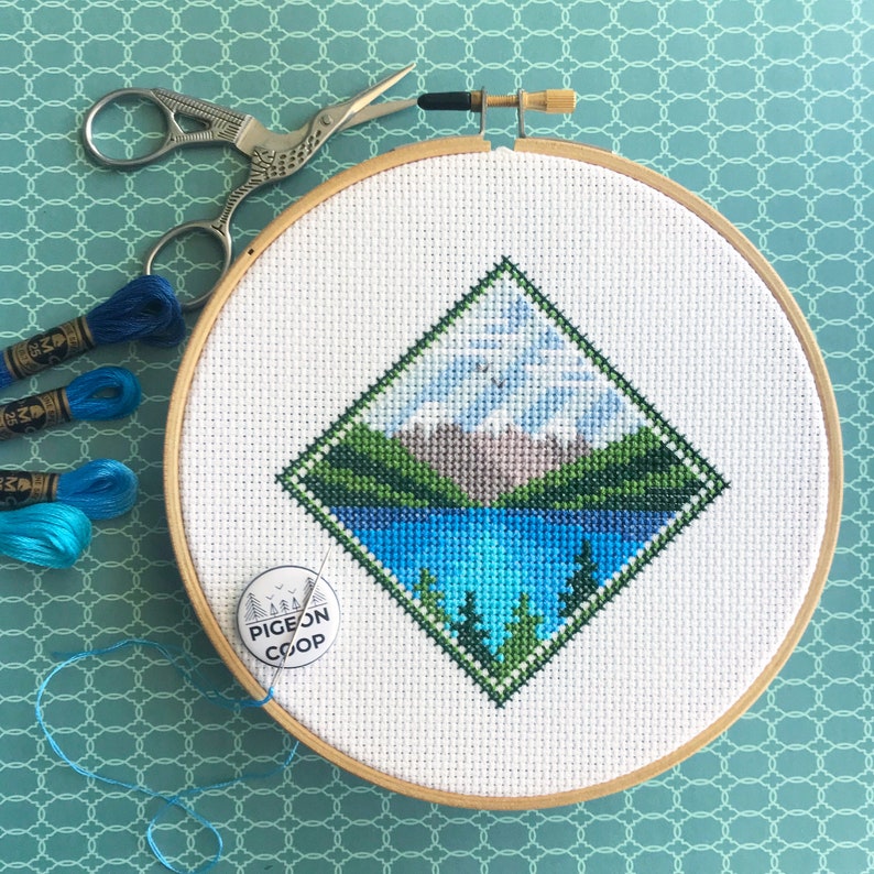 May include: A completed cross-stitch embroidery hoop with a diamond-shaped design of a mountain lake scene. The design features a blue lake, green trees, and a mountain range with a light blue sky. The hoop is made of wood and has a white fabric background. The embroidery is framed with a green border. A small round sticker with the text "PIGEON COOP" is visible in the lower right corner of the hoop.