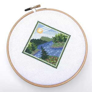 Puede incluir: Un aro de bordado de punto de cruz con una escena de paisaje de un lago, &aacute;rboles y un sol. La escena est&aacute; bordada en tonos de verde, azul y amarillo.