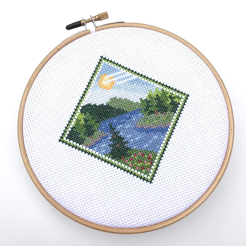 Puede incluir: Un aro de bordado de punto de cruz con una escena de paisaje de un lago, &aacute;rboles y un sol. La escena est&aacute; bordada en tonos de verde, azul y amarillo.